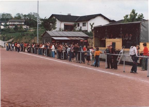 Menschenmenge steht an einer Kasse neben einem Sportplatz, im Hintergrund sind Geb&auml;ude und B&auml;ume sichtbar.