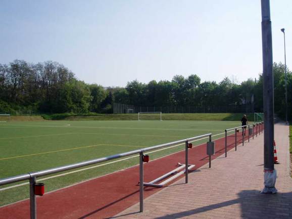 Leeres Fu&szlig;ballfeld mit Kunstrasen, umrahmt von einem Zaun und B&auml;umen im Hintergrund, eine Person geht entlang des Weges.