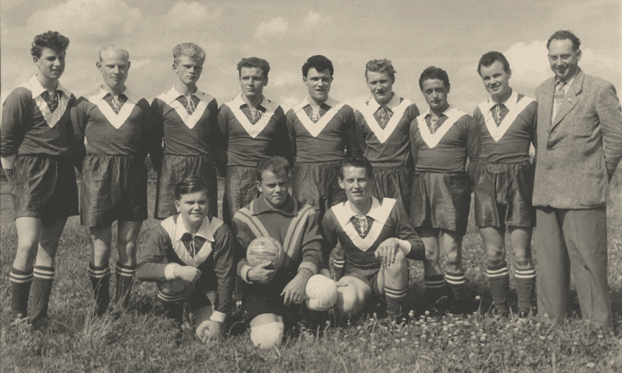 Schwarz-wei&szlig;-Foto einer Fu&szlig;ballmannschaft mit zw&ouml;lf Spielern in einheitlichen Trikots und einem Trainer im Hintergrund.