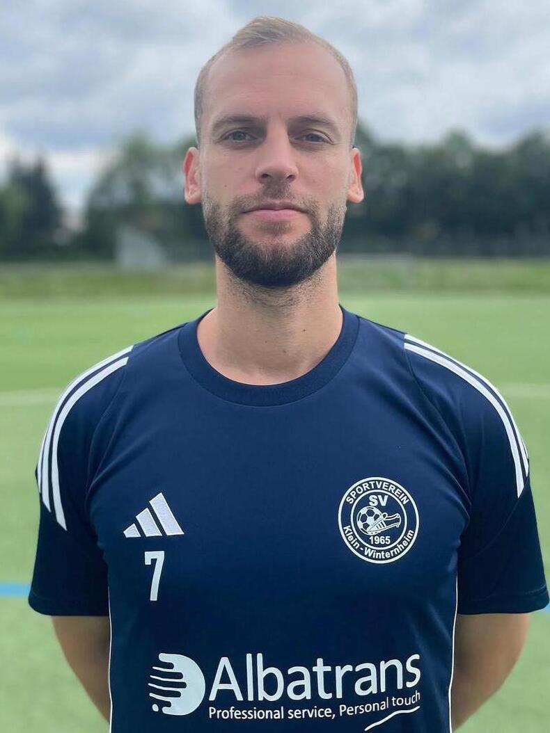 Portrait eines Mannes in einem blauen Sporttrikot mit Vereinslogo, stehend auf einem Fu&szlig;ballfeld.