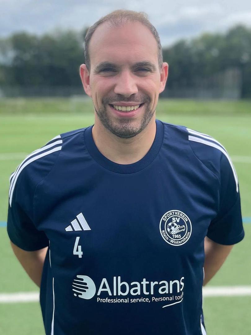 L&auml;chelnder Mann in dunkelblauem Fu&szlig;balltrikot mit Vereinslogo und Sponsor auf einem Sportplatz hinter ihm.