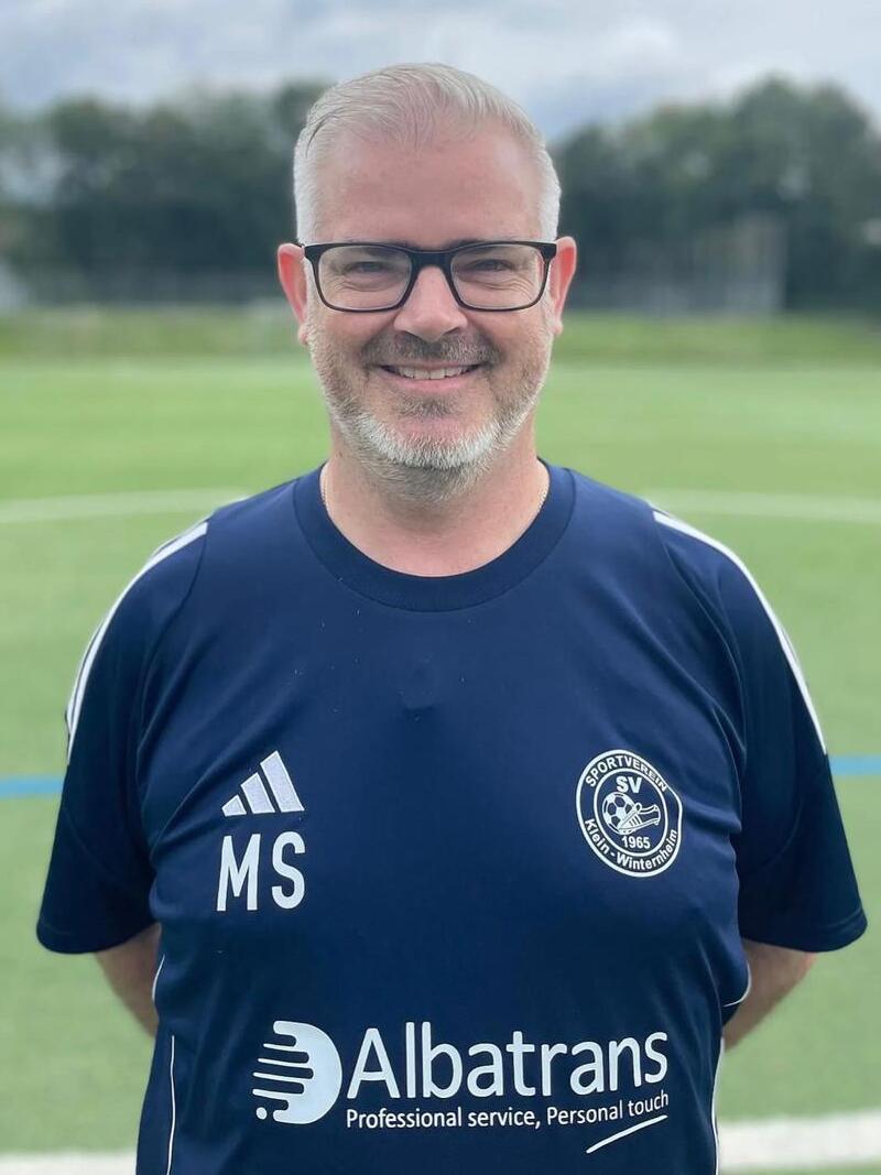 Portrait eines l&auml;chelnden Mannes in einem blauen Fu&szlig;balltrikot mit Logo und Sponsorenaufdruck auf einem Sportplatz.