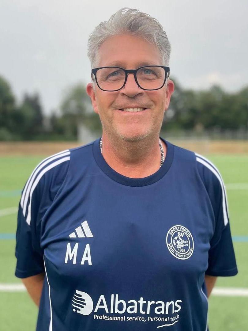 Mann mit grauem Haar und Brille, tr&auml;gt ein dunkelblaues Fu&szlig;balltrikot auf einem Sportplatz.