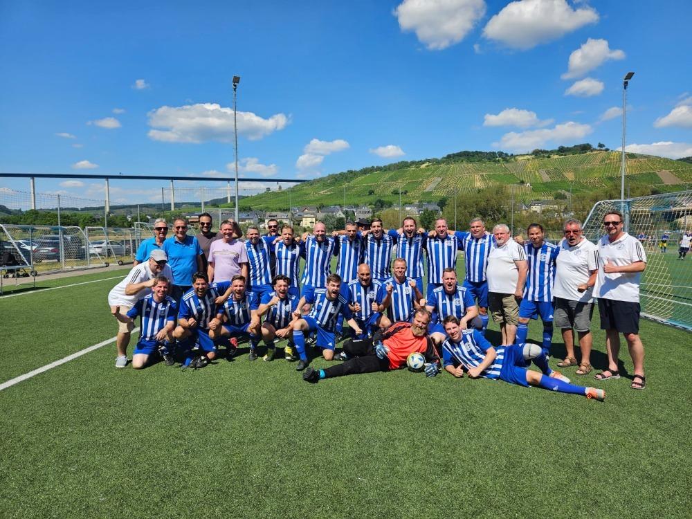 Gruppenbild von Fu&szlig;ballspielern in blauen Trikots mit wei&szlig;en Streifen auf einem Sportplatz vor einer h&uuml;geligen Landschaft.