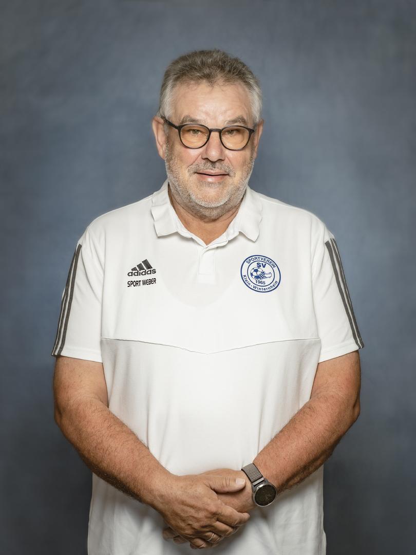 Portr&auml;t eines &auml;lteren Mannes mit Brille, in einem wei&szlig;en Sportshirt mit Vereinslogo, die Arme vor der Brust verschr&auml;nkt.