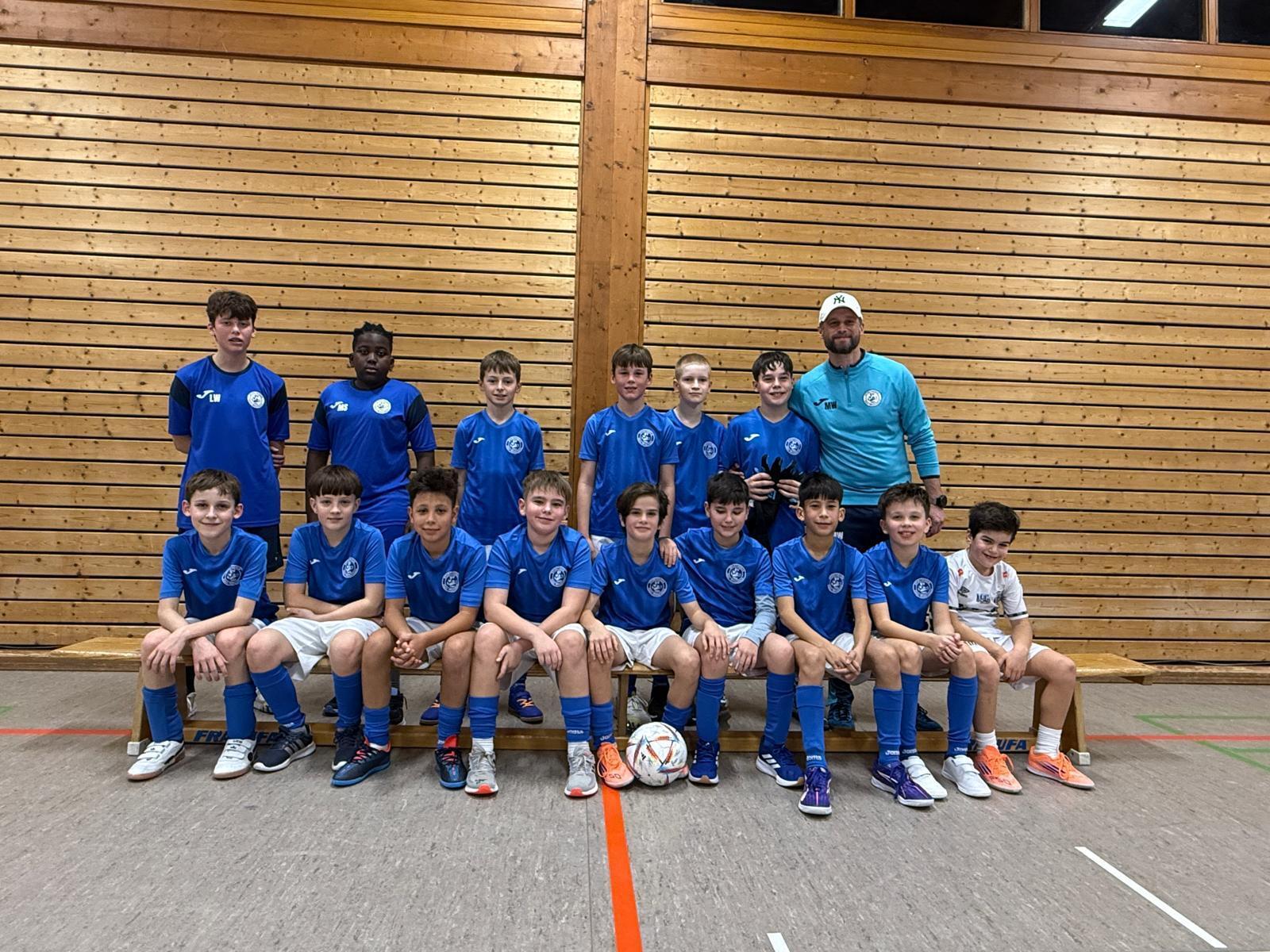 Gruppenfoto einer Fu&szlig;ballmannschaft von Jungen in blauen Trikots, mit Trainer, in einer Sporthalle vor Holzpaneelen.