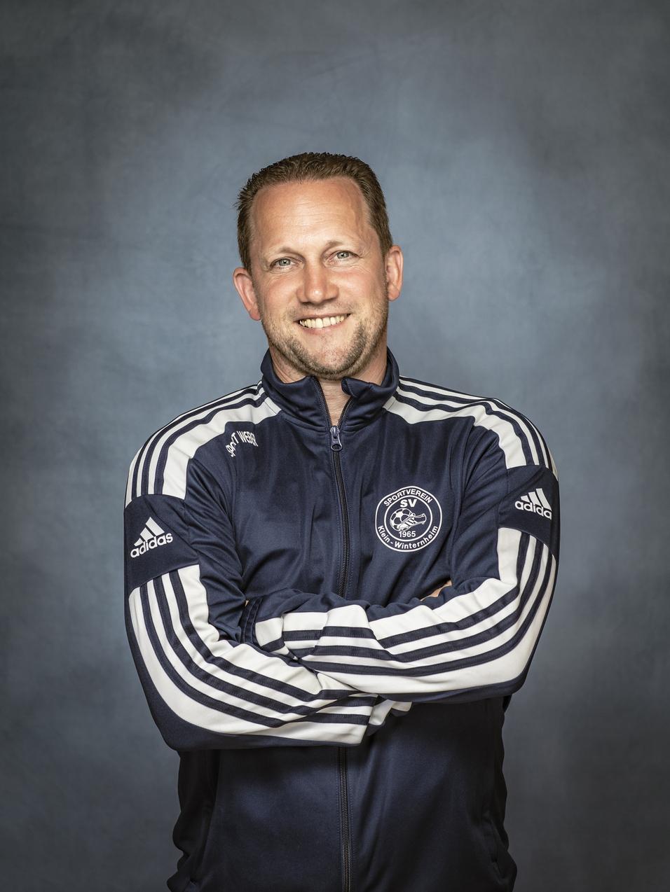 Mann in Sportjacke mit geschlossenen Armen, l&auml;chelt vor einem neutralen Hintergrund.