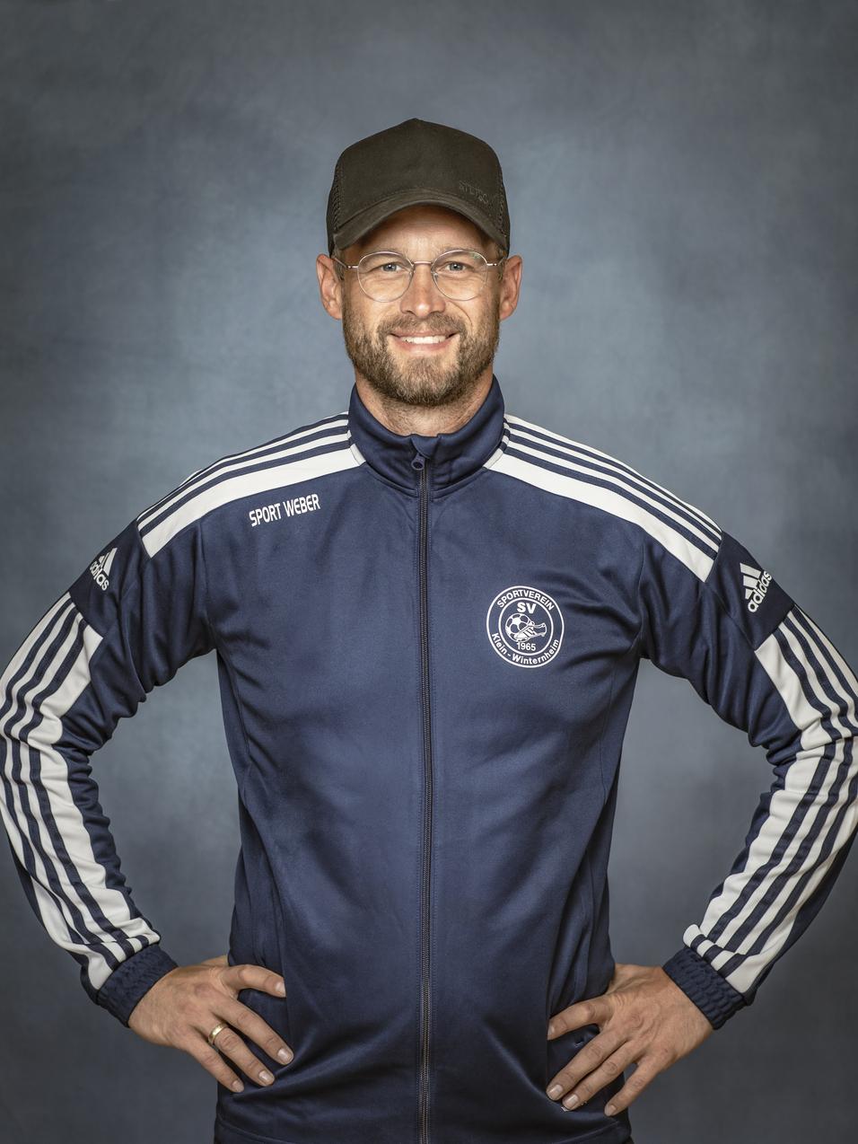 Portr&auml;t eines Mannes in sportlicher Kleidung mit Brille und M&uuml;tze, l&auml;chelnd vor einem neutralen Hintergrund.