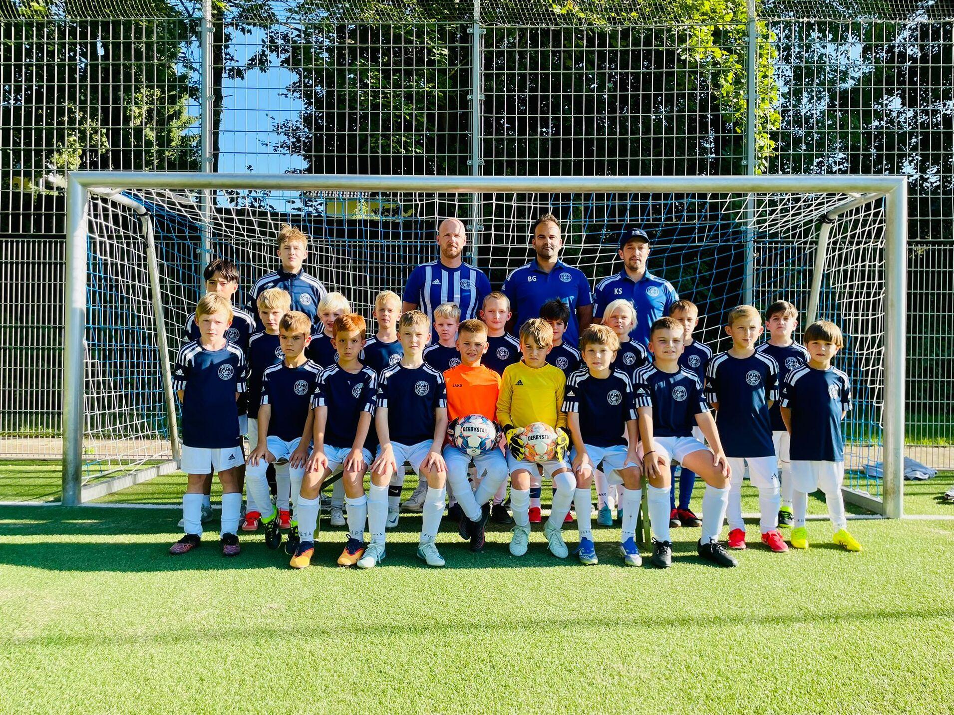 Gruppe von jugendlichen Fu&szlig;ballspielern in blauen Trikots mit Trainern vor einem Tor auf einem Kunstrasenplatz.