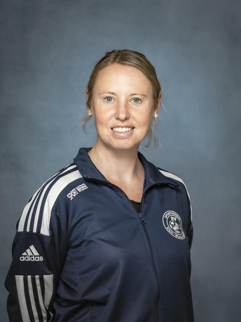 Portr&auml;t einer Frau in einem blauen Sportjackett mit Vereinslogo, l&auml;chelnd vor einem graublauen Hintergrund.