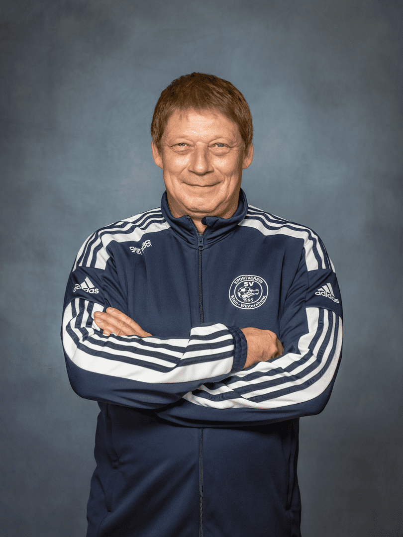 Portr&auml;t eines l&auml;chelnden Mannes mit kurzen Haaren in einer blauen Sportjacke mit wei&szlig;en Streifen und verschr&auml;nkten Armen.