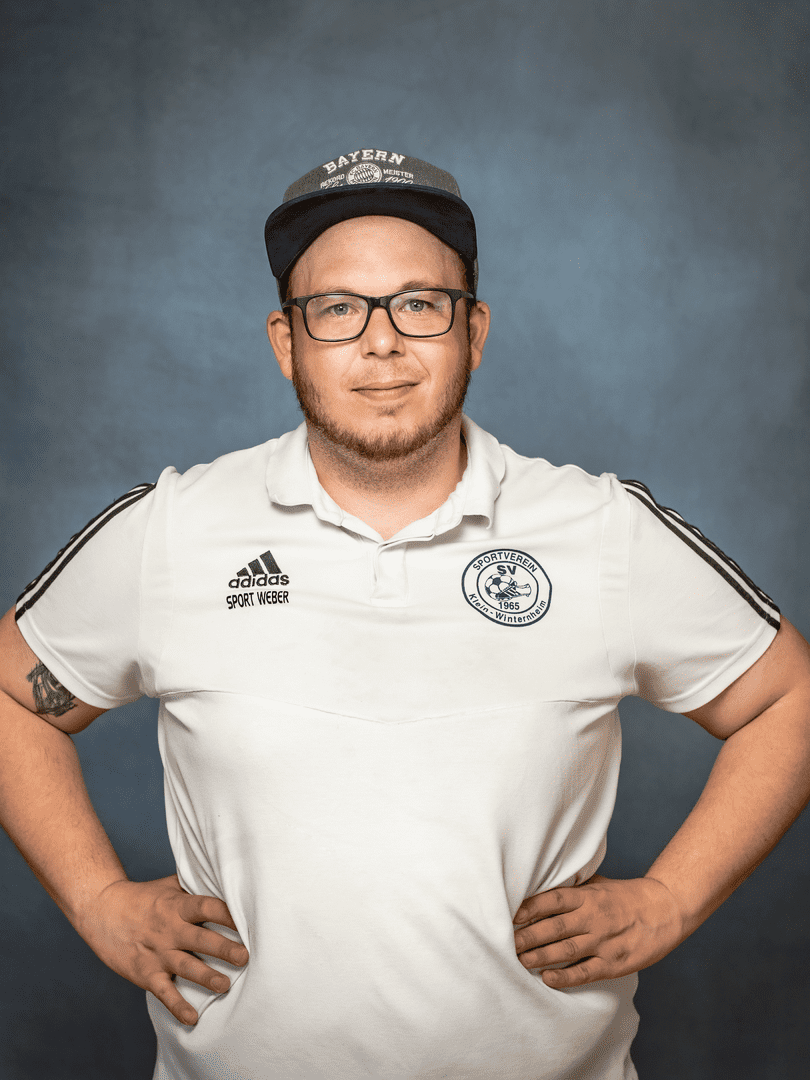 Portr&auml;t eines Mannes mit kurzem Bart, Brille und einem Kapit&auml;nshut, der in einem wei&szlig;en Sportshirt mit Logo posiert.