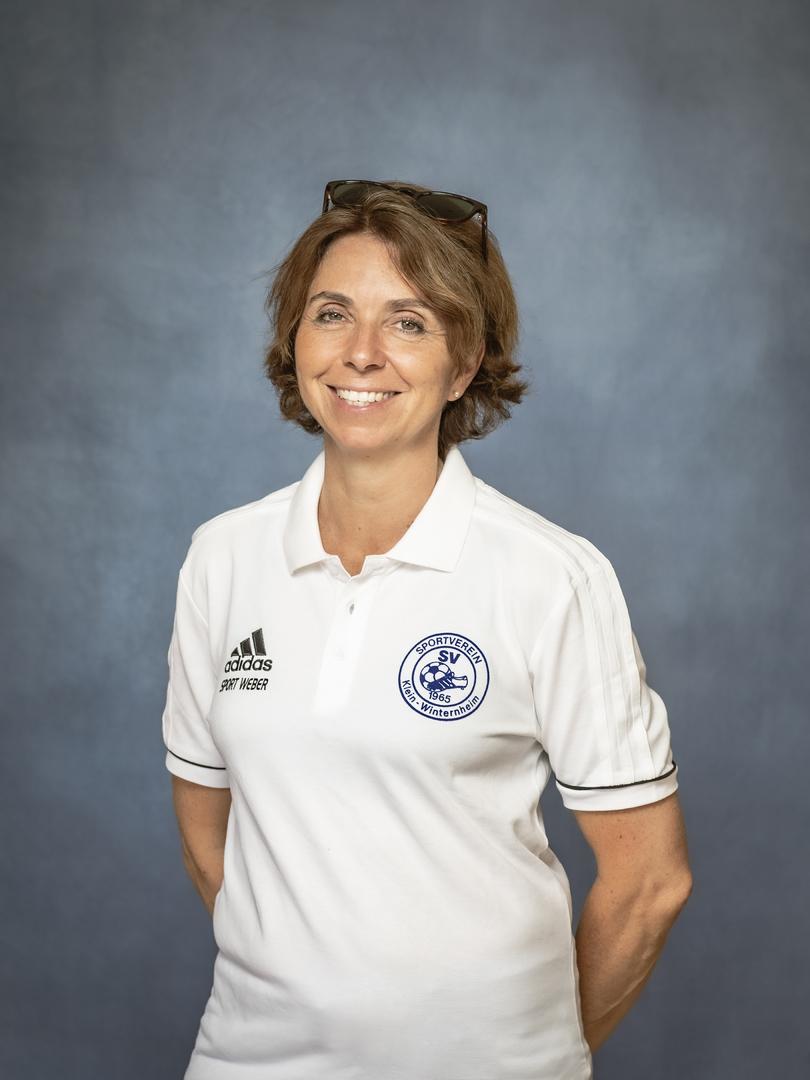 Portrait einer l&auml;chelnden Frau in einem wei&szlig;en Polohemd mit Vereinslogo und Sonnenbrille im Haar, vor grauem Hintergrund.