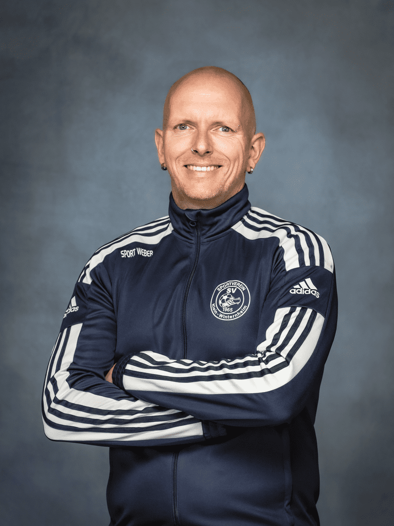 Portrait eines l&auml;chelnden Mannes in Sportjacke mit Logo, Arme vor der Brust verschr&auml;nkt, vor neutralem Hintergrund.