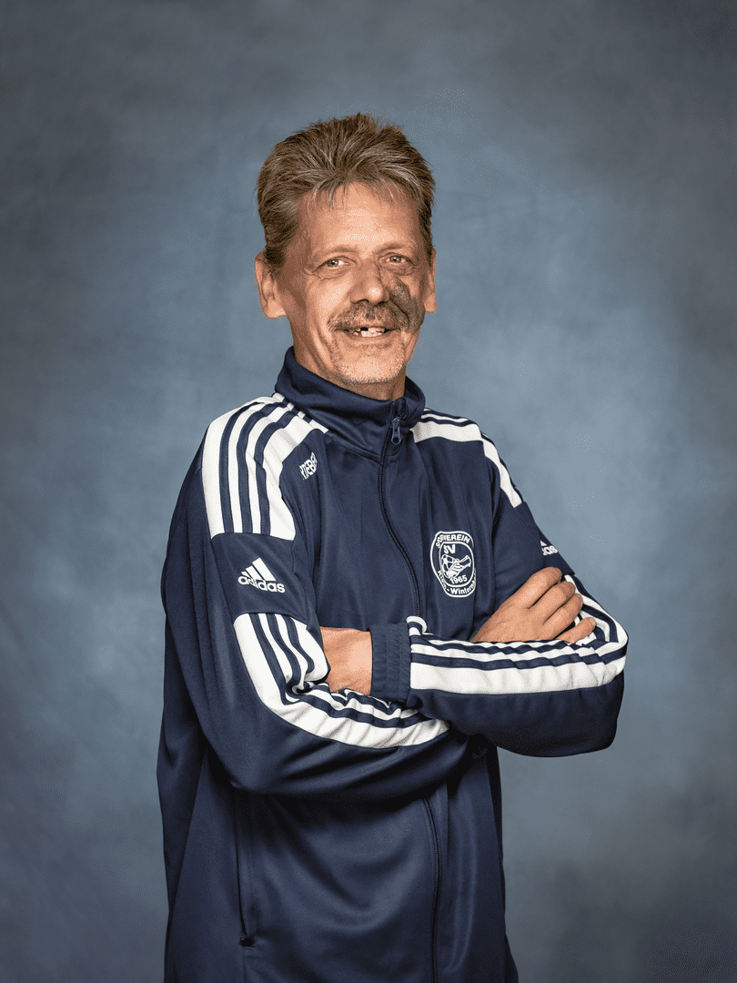 Mann mit grauen Haaren in sportlicher Adidas-Jacke, die Arme vor der Brust verschr&auml;nkt, l&auml;chelt in die Kamera.