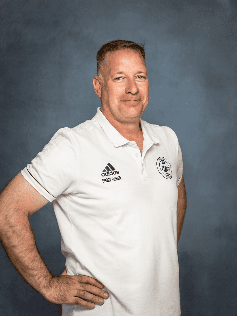 Portr&auml;t eines l&auml;chelnden Mannes in einem wei&szlig;en Poloshirt vor einem blauen Hintergrund, die H&auml;nde an der H&uuml;fte.