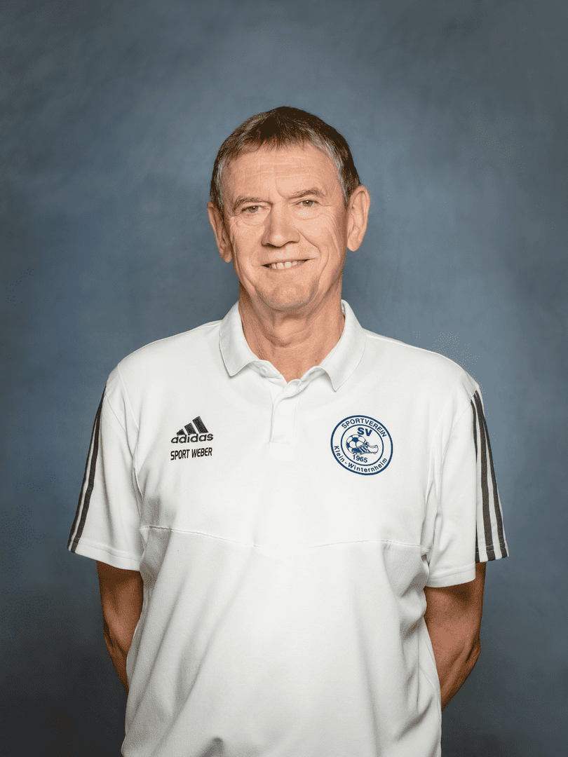 Portrait eines Mannes in einem wei&szlig;en Polo mit Vereinslogo und Adidas-Streifen, vor einem graublauen Hintergrund.