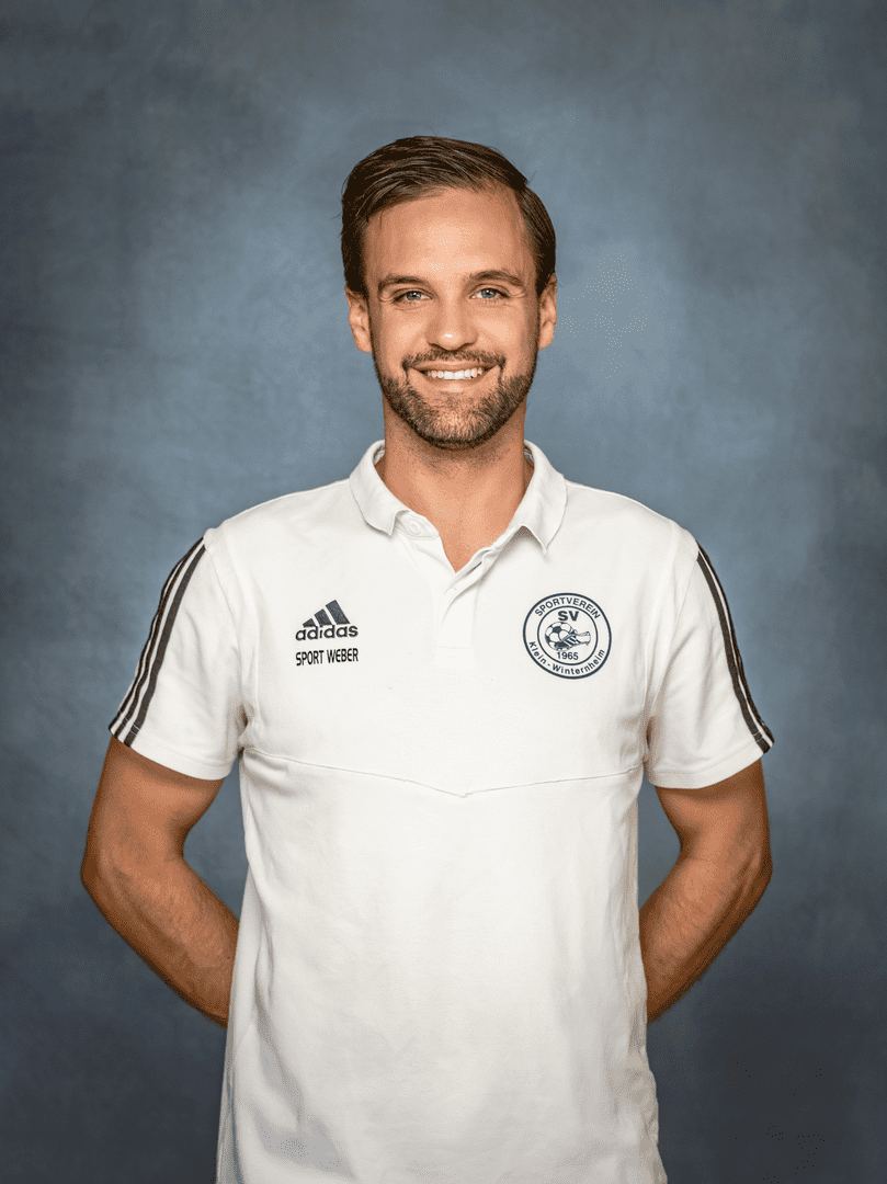 Portr&auml;t eines l&auml;chelnden Mannes in wei&szlig;em Poloshirt mit Vereinslogo und adidas-Logo vor grauem Hintergrund.