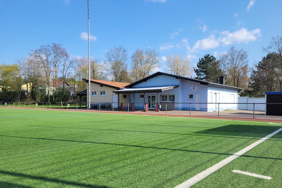 Sportplatz mit Kunstrasen und Vereinsgeb&auml;ude im Hintergrund, umgeben von B&auml;umen und unter blauem Himmel.