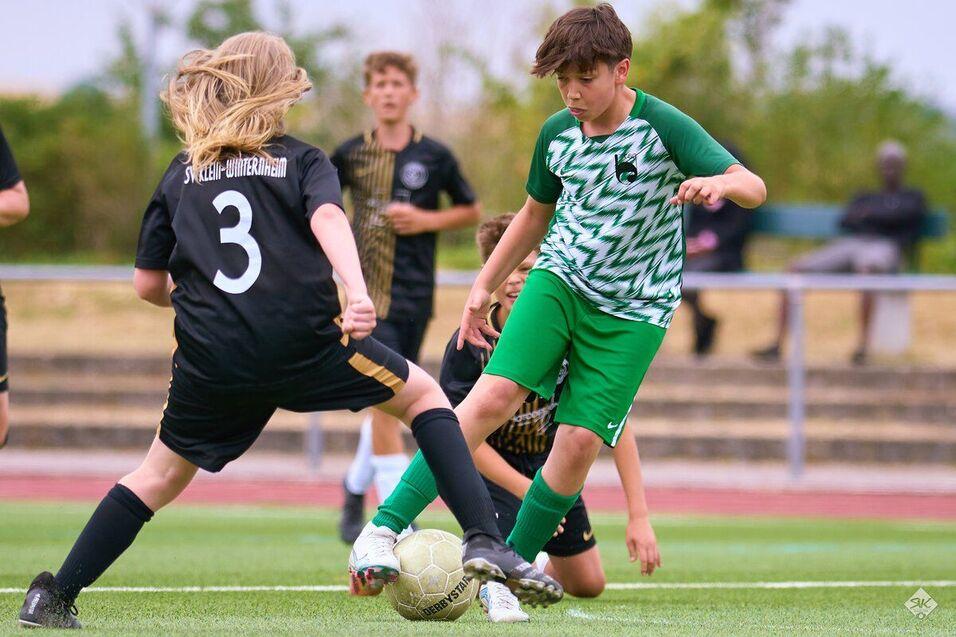 Zwei Kinder spielen Fu&szlig;ball, w&auml;hrend ein Spieler versucht, den Ball zu erobern. Sie tragen gr&uuml;ne und schwarze Trikots.