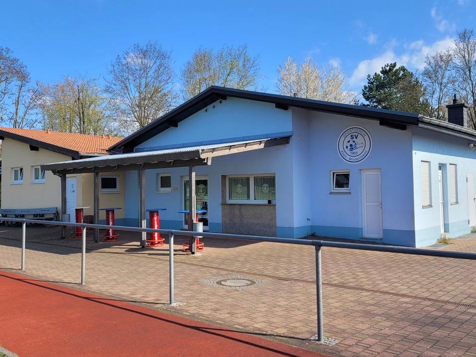 Vorderansicht eines blauen Vereinsgeb&auml;udes mit Parkplatz und Sportplatz im Hintergrund an einem sonnigen Tag.